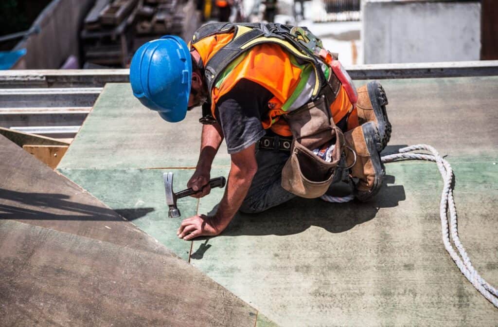 Tradie on a roof using a hammer. There are a number of ways seeing a podiatrist can help with foot and leg pain in tradies.